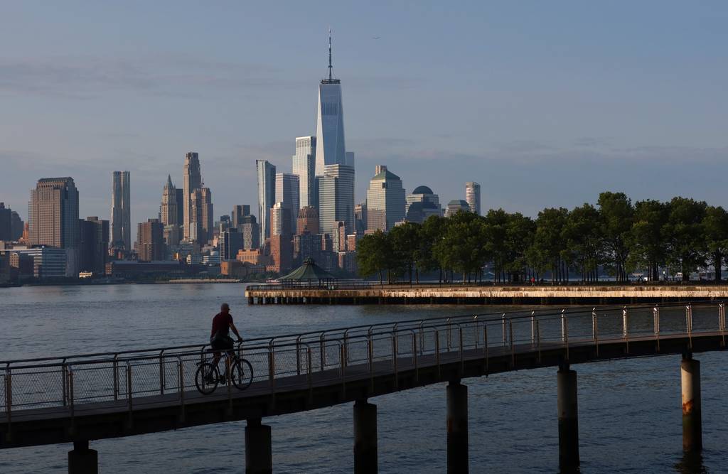 The sun rises on the skyline of lower Manhattan and One World Trade Center in New York City as a person rides a bicycle on a pier in the Hudson River on June 9, 2023, in Hoboken, New Jersey. 